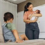 A mother lovingly cradles her baby while her child sits nearby on the couch in a modern room setting.