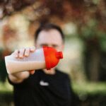 Focus on a man holding a plastic protein shake tumbler outdoors with a blurred background.