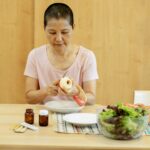 Middle aged ethnic female with short hair in t shirt peeling fresh apple and preparing salad while sitting at table with veggies and various medicine during remission