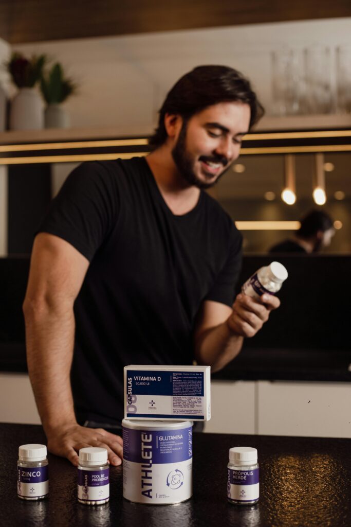 Adult man standing in a kitchen examining a vitamin bottle with more supplements on the countertop.